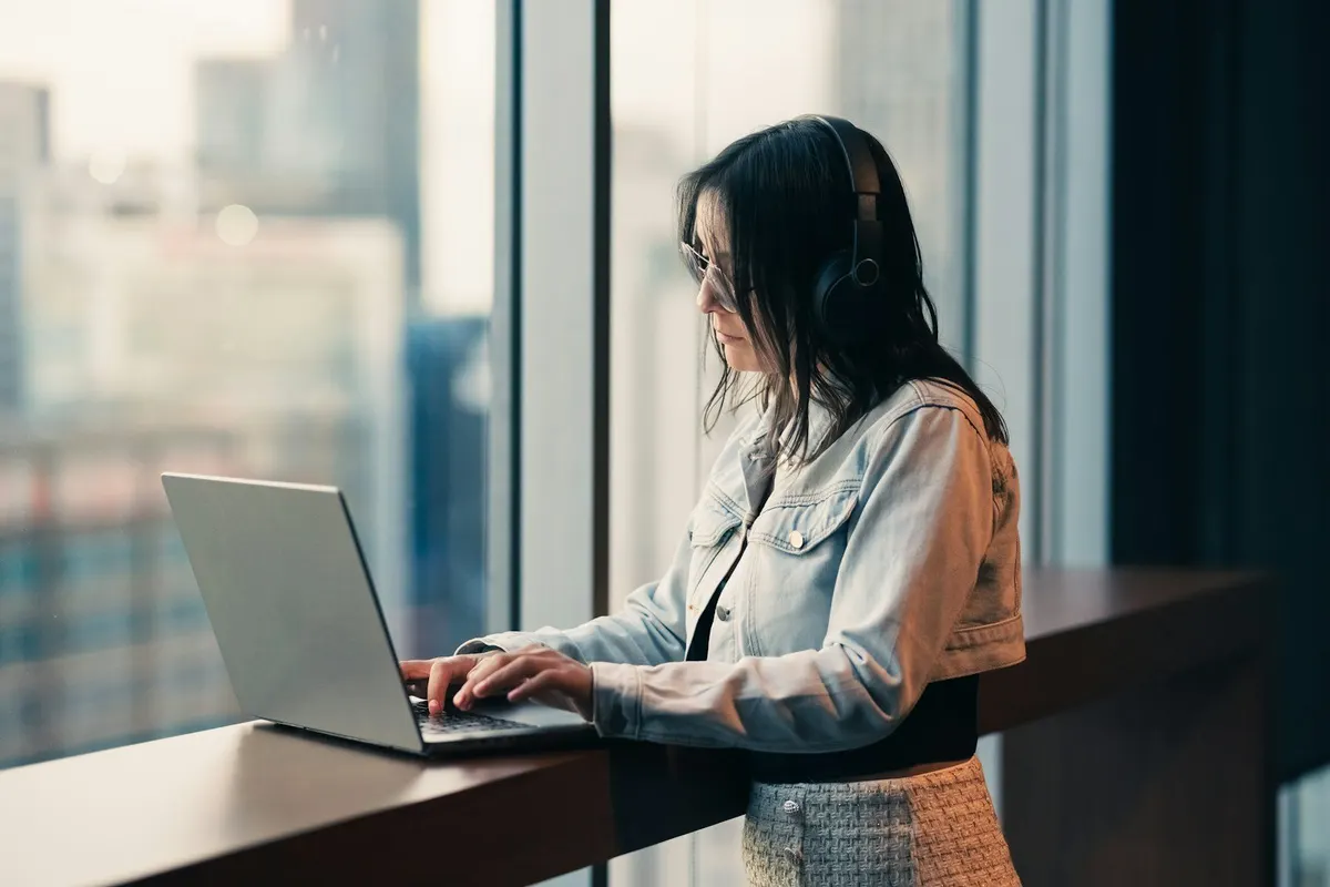 Person wearing headphones working on a laptop by a large window.